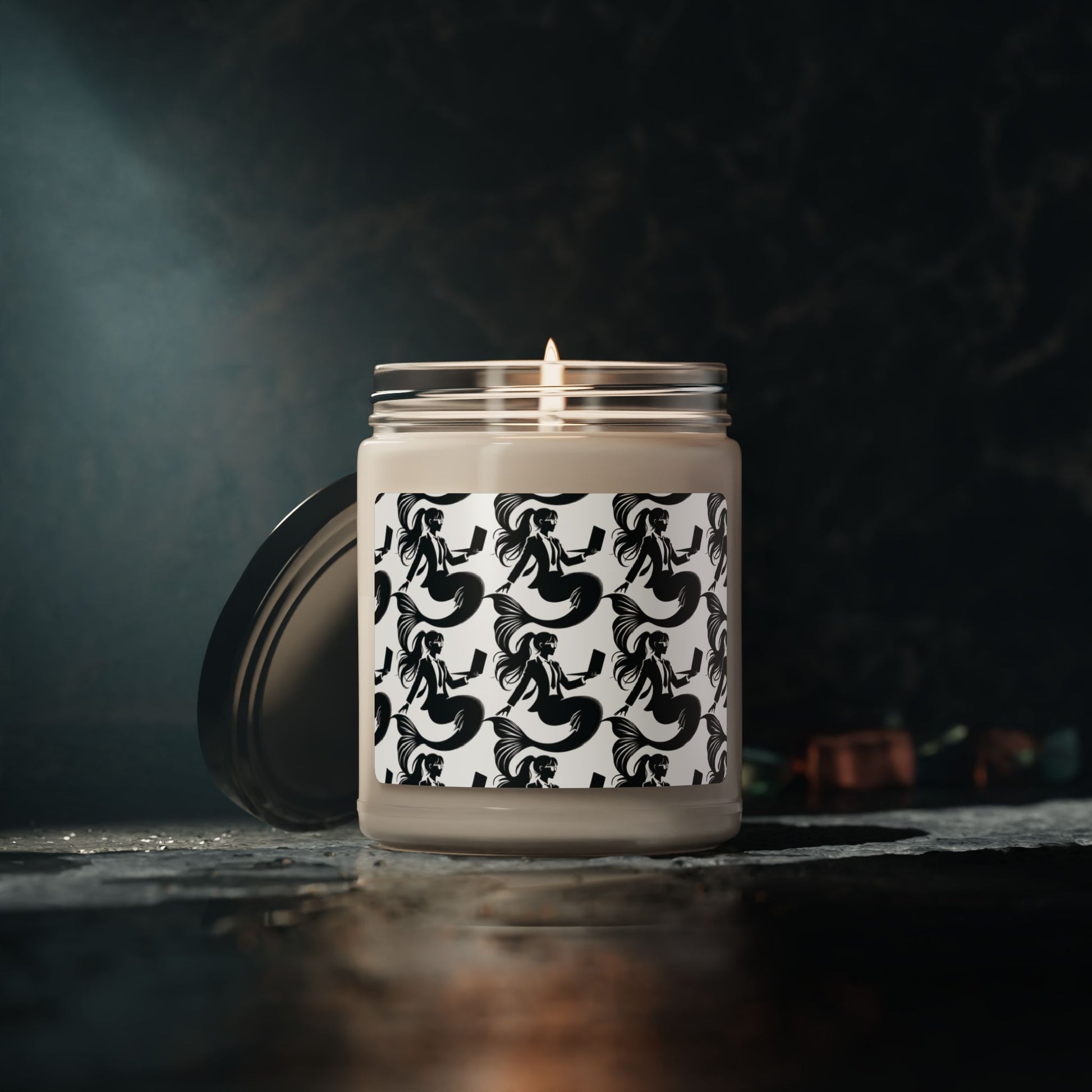 Corporate mermaids on a soy candle glass jar with black lid laying off and against the jar. The corporate mermaid design is a black and white mermaid wearing a suit, glasses and hair in a ponytail, holding an open laptop. 