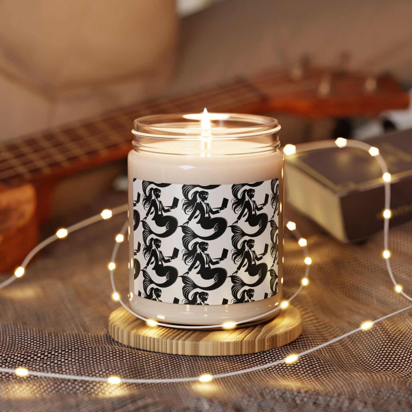 Corporate mermaids on a soy candle glass jar without a lid, wrapped in string lights, sitting on a wooden coaster in front of a guitar. The corporate mermaid design is a black and white mermaid wearing a suit, glasses and hair in a ponytail, holding an open laptop. 