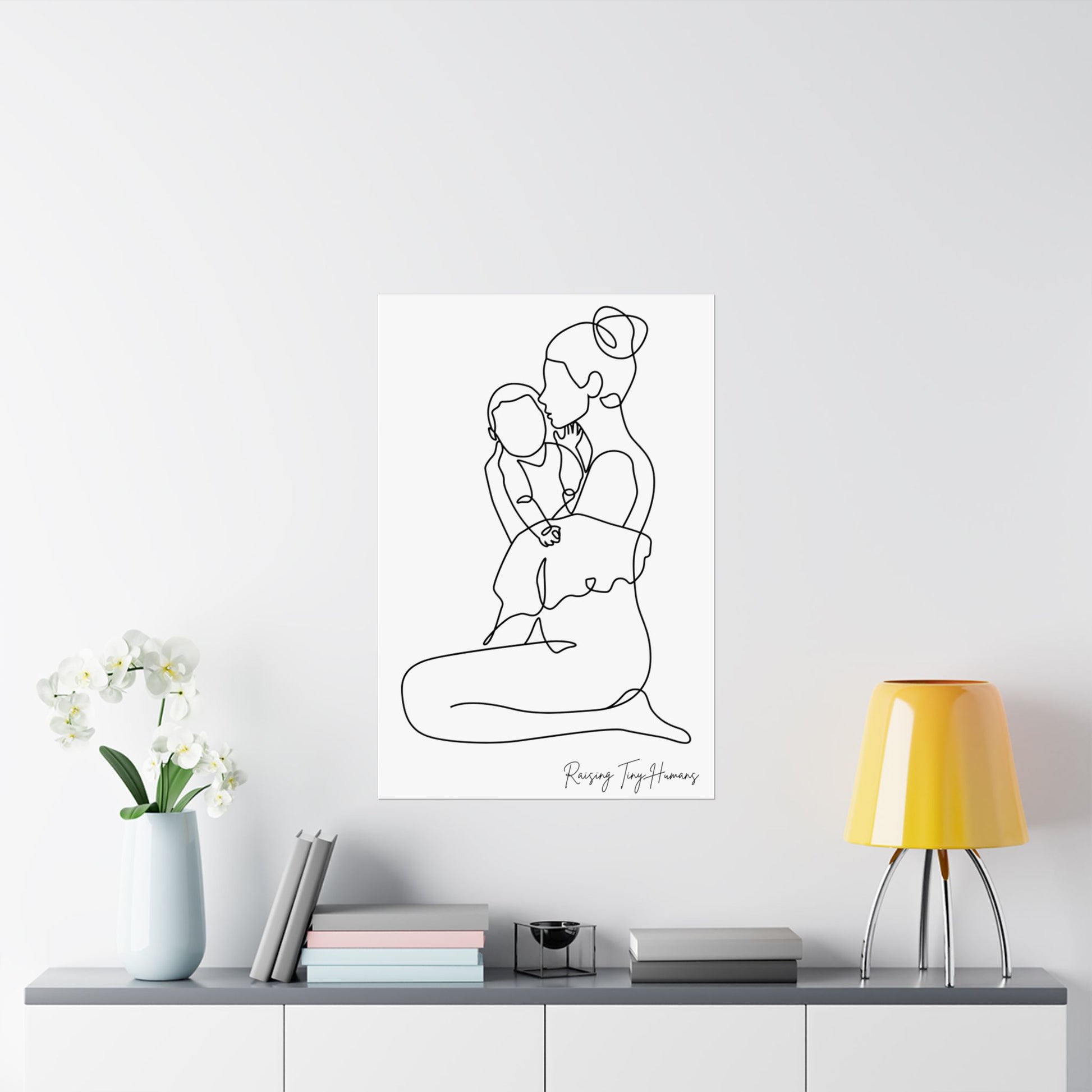 White poster hung on a wall above a credenza holding flower pot and books, a lamp and other accessories. The poster is a line drawing of mom holding a baby and the words "raising tiny humans" in handwriting place at the bottom right corner