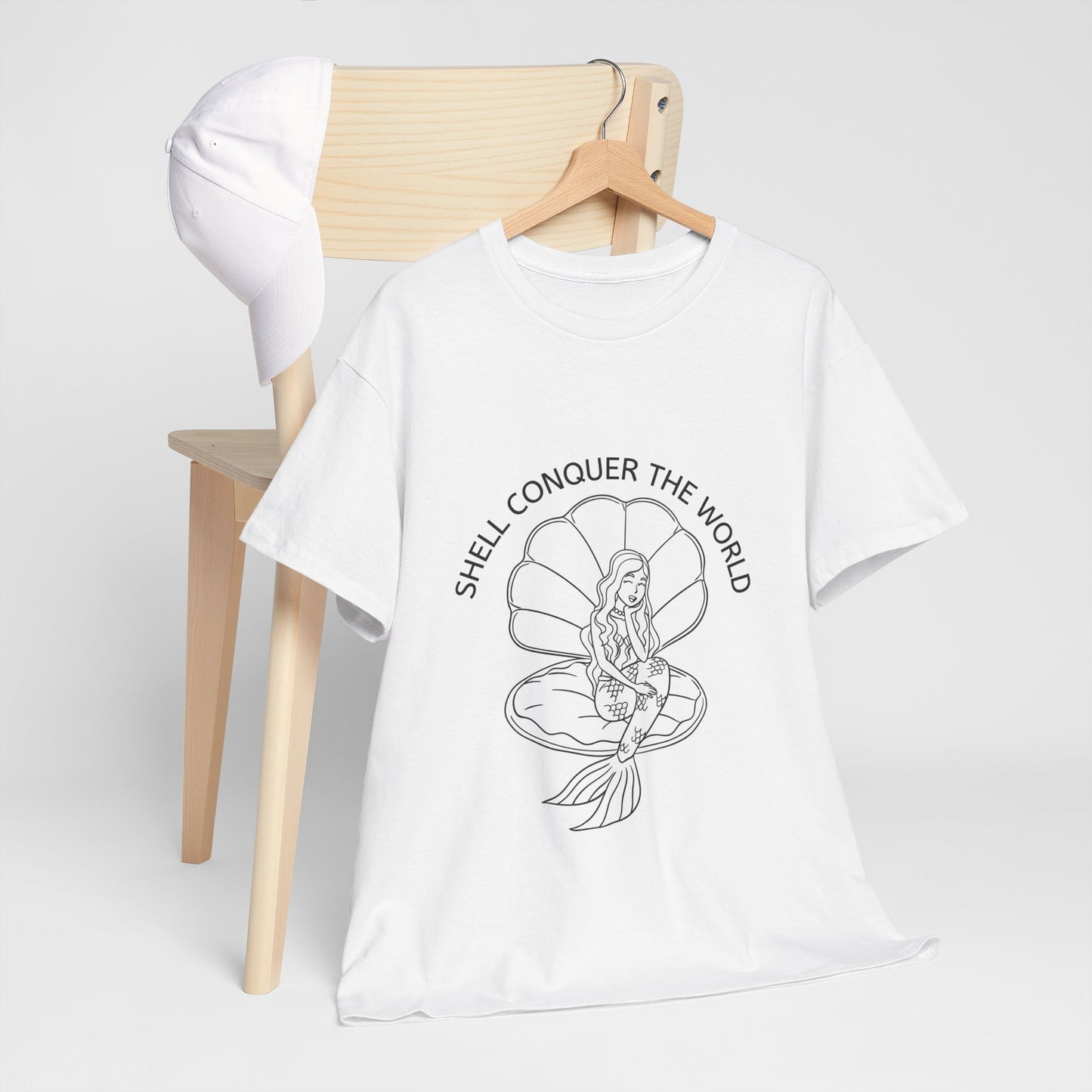 White unisex short sleeve t-shirt on a hanger, hanging on a chair with a plain white hat hanging on the other side. The white t-shirt has a design of a mermaid sitting in an open oyster shell, with the words "SHELL CONQUER THE WORLD" written in an arc above the image. 